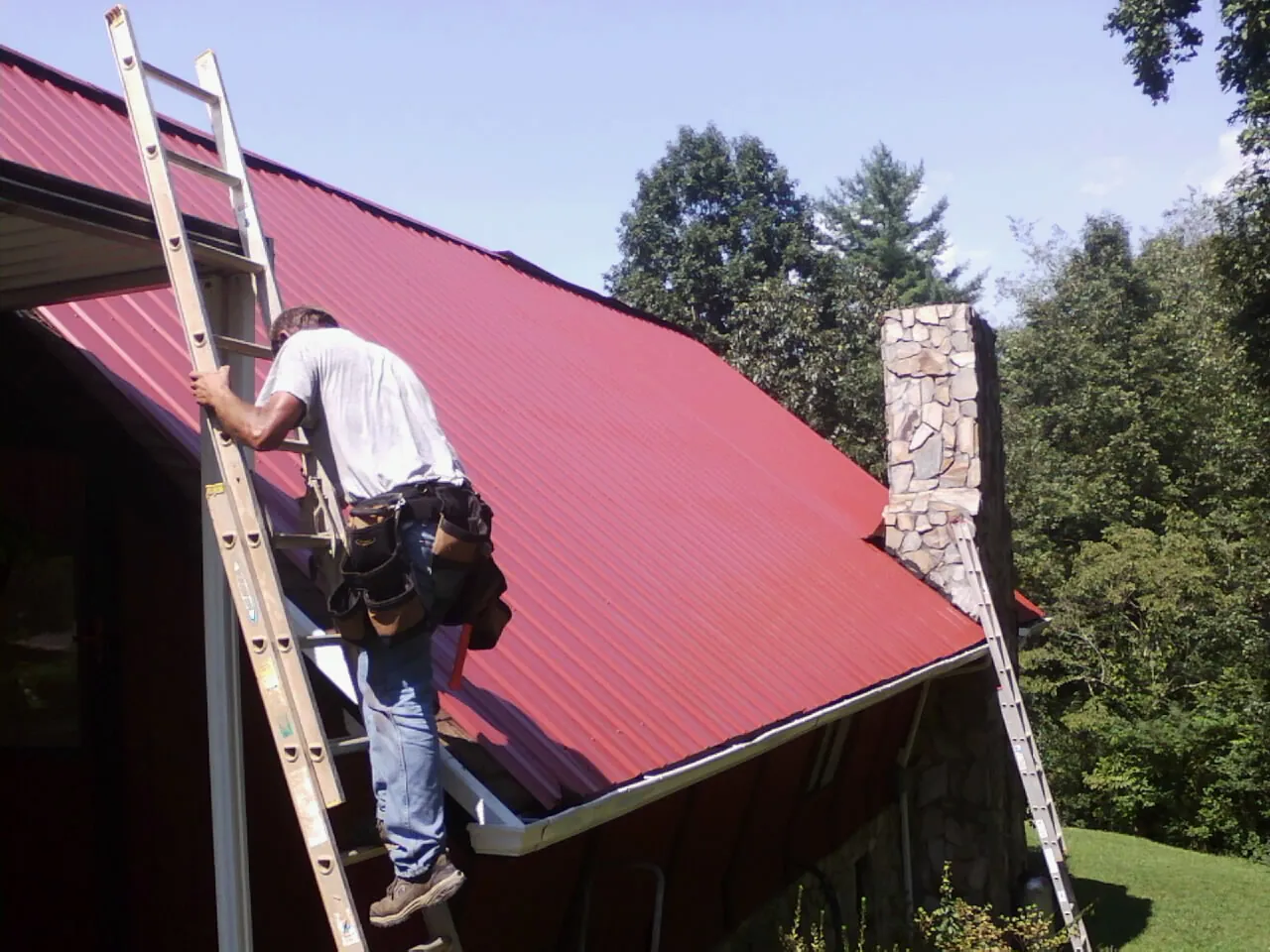 Roofer on a ladder beside a red metal roof and stone chimney — Soffit & Fascia Repair by Roofing Brothers in Thorp